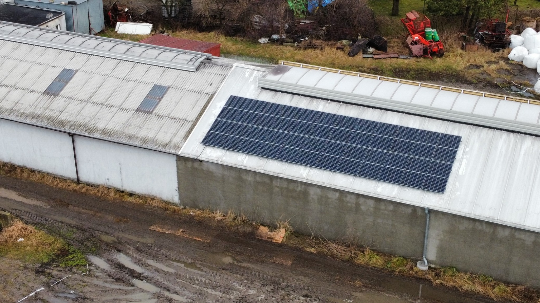 Instalacja paneli fotowoltaicznych na dachu budynku gospodarczego z widocznymi oknami dachowymi i błotnistym terenem wokół.