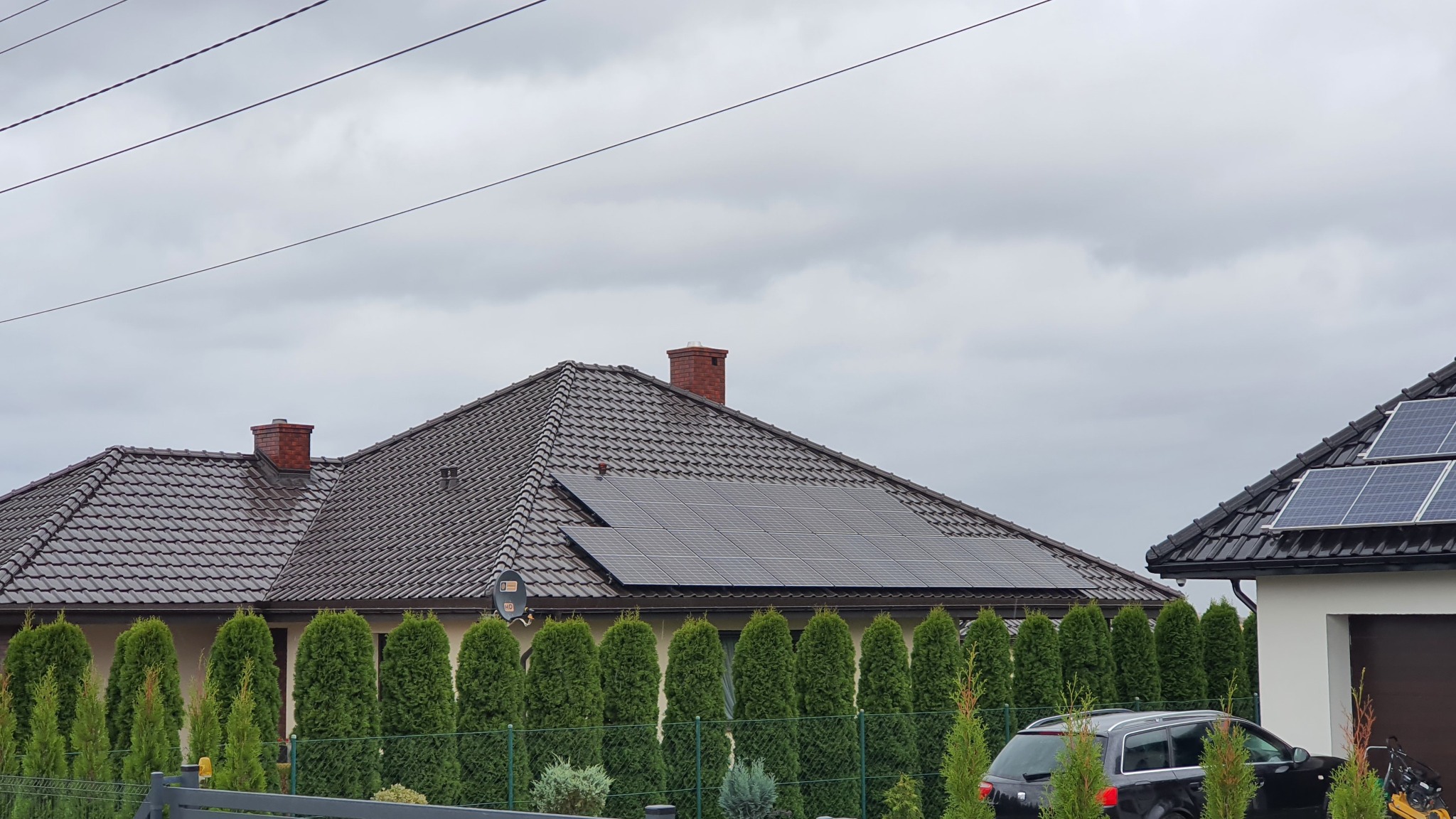 Dom jednorodzinny z brązową dachówką i panelami fotowoltaicznymi na dachu, widok z poziomu ulicy w pochmurny dzień, z zielonymi tujami wzdłuż ogrodzenia.