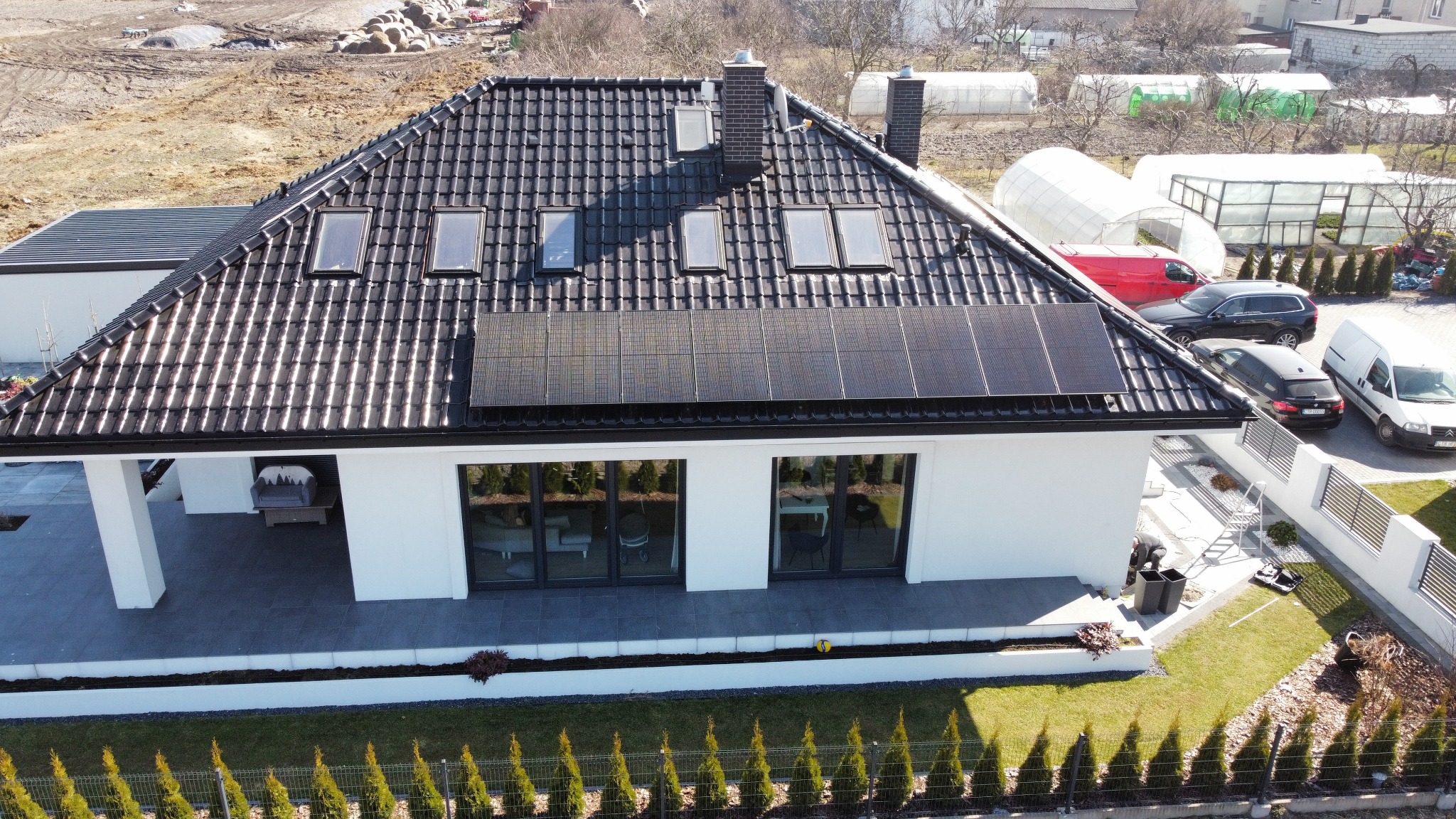 Widok z góry na nowoczesny dom z ciemną dachówką, oknami dachowymi i panelami słonecznymi, otoczony zadbanym ogrodem z tujami i szarym tarasem. W tle widoczne szklarnie i zaparkowane samochody.
