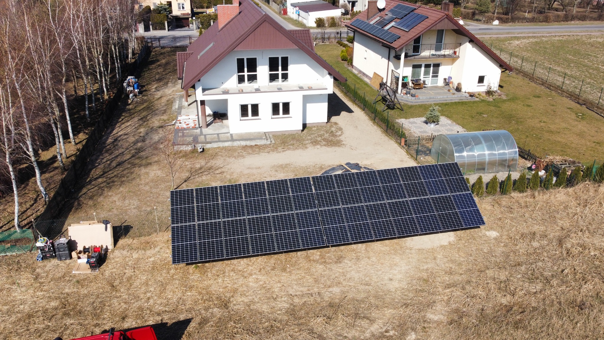 Widok z drona na rozłożone panele fotowoltaiczne na trawniku przed domem jednorodzinnym z panelami na dachu, w tle szklarnia i sprzęt montażowy.