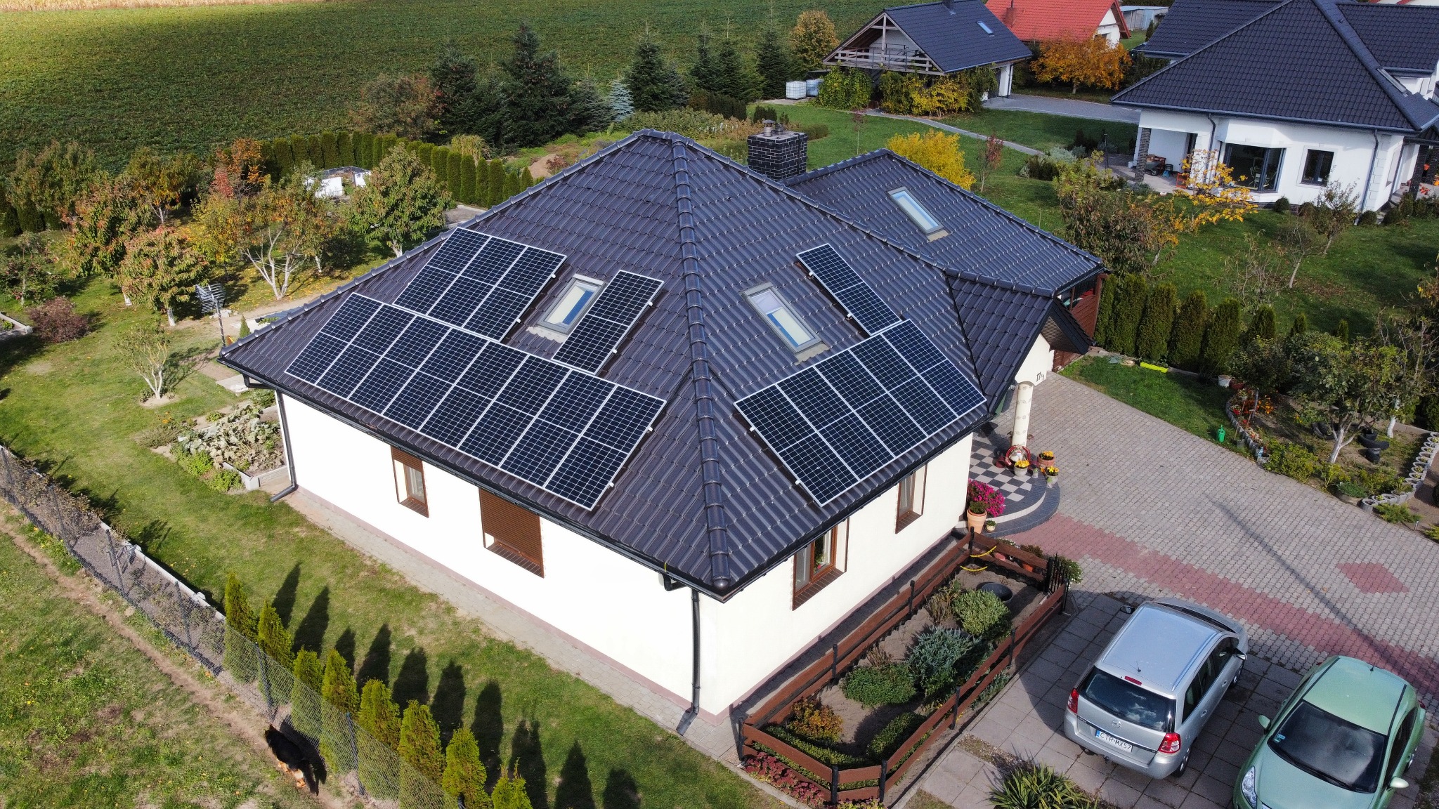 Dom jednorodzinny z ciemną dachówką i panelami fotowoltaicznymi zamontowanymi na dachu, widok z lotu ptaka w słoneczny dzień. Na podjeździe zaparkowane dwa samochody.