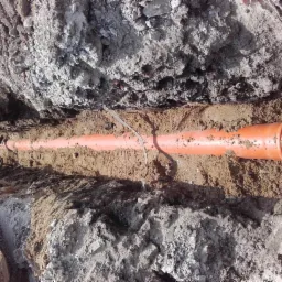 Pomarańczowa rura kanalizacyjna ułożona w wykopie ziemnym z widocznymi śladami po pracy koparki i luźną taśmą ostrzegawczą.