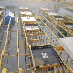 Szeroki kadr z góry na plac budowy z licznymi szalunkami pod fundamenty, wspartymi na regulowanych podporach, z widocznymi fragmentami zbrojenia i małym niebieskim barakiem w tle.