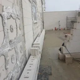 Ściana budynku w trakcie budowy z betonu komórkowego, widoczne bloczki ułożone wzdłuż ściany, ślady zaprawy z dekoracyjnymi elementami, okno, oraz narzędzia i materiały budowlane w tle.