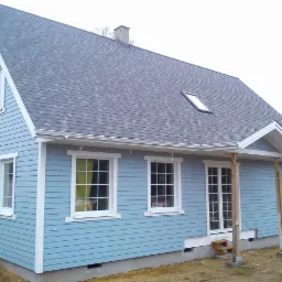 Niebieski dom z szarym dachem i białymi oknami, z widoczną fakturą desek na elewacji. Widoczne schodki przed wejściem i fragment terenu wok&oacute;ł domu.