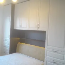 Biała szafa na wymiar z szufladami i szafkami wiszącymi nad łóżkiem. Widoczne metalowe uchwyty i zagłówek łóżka. Całość w jasnym, minimalistycznym stylu.