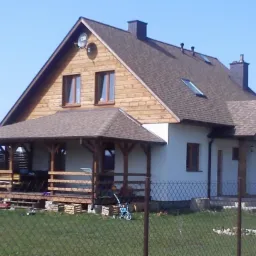 Dwukondygnacyjny dom z brązowym dachem i drewnianą elewacją na poddaszu. Zadaszony drewniany taras z balustradą. Widoczne zabawki na trawniku, ogrodzenie.