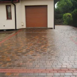 Nowo ułożony, wilgotny podjazd z kostki brukowej w odcieniach brązu i czerwieni, obramowany czerwoną kostką, prowadzący do garażu w jasnym budynku z brązową bramą.