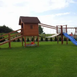 Drewniany plac zabaw z domkiem, zjeżdżalnią i huśtawką na zielonym trawniku, otoczony tujami. Dzieci bawią się na zjeżdżalni i huśtawce. Słoneczny dzień.