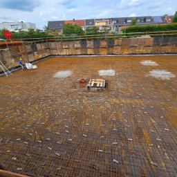 Firma Ogólnobudowlana Reno Piotr Wiśniewski - Szeroki wykop zabezpieczony metalowymi panelami, z ułożoną siatką zbrojeniową na czarnej folii, widoczne podpory i pracownik w kasku z workiem, częściowo pochmurne niebo.