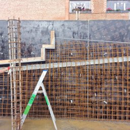 Firma Ogólnobudowlana Reno Piotr Wiśniewski - Pracownik w kasku i kombinezonie, z czerwoną rękawicą, przy zbrojeniu pionowego słupa na tle siatki zbrojeniowej ściany i drabiny aluminiowej z zielonymi elementami, widok na budynek z cegły...