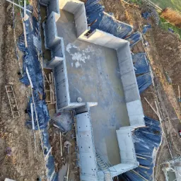 Widok z góry na fundamenty domu w trakcie budowy, z widoczną hydroizolacją z czarnej folii, deskowaniem i niewielką ilością wody wewnątrz, na tle ziemi i zieleni.