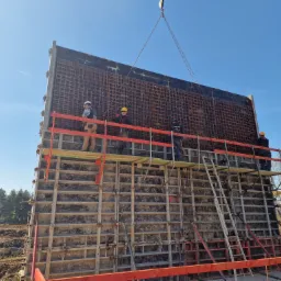 Montaż szalunku zbrojeniowego na budowie, pracownicy w kaskach na rusztowaniu, widoczne stalowe siatki zbrojeniowe i drewniane elementy konstrukcyjne, w tle błękitne niebo i drzewa.