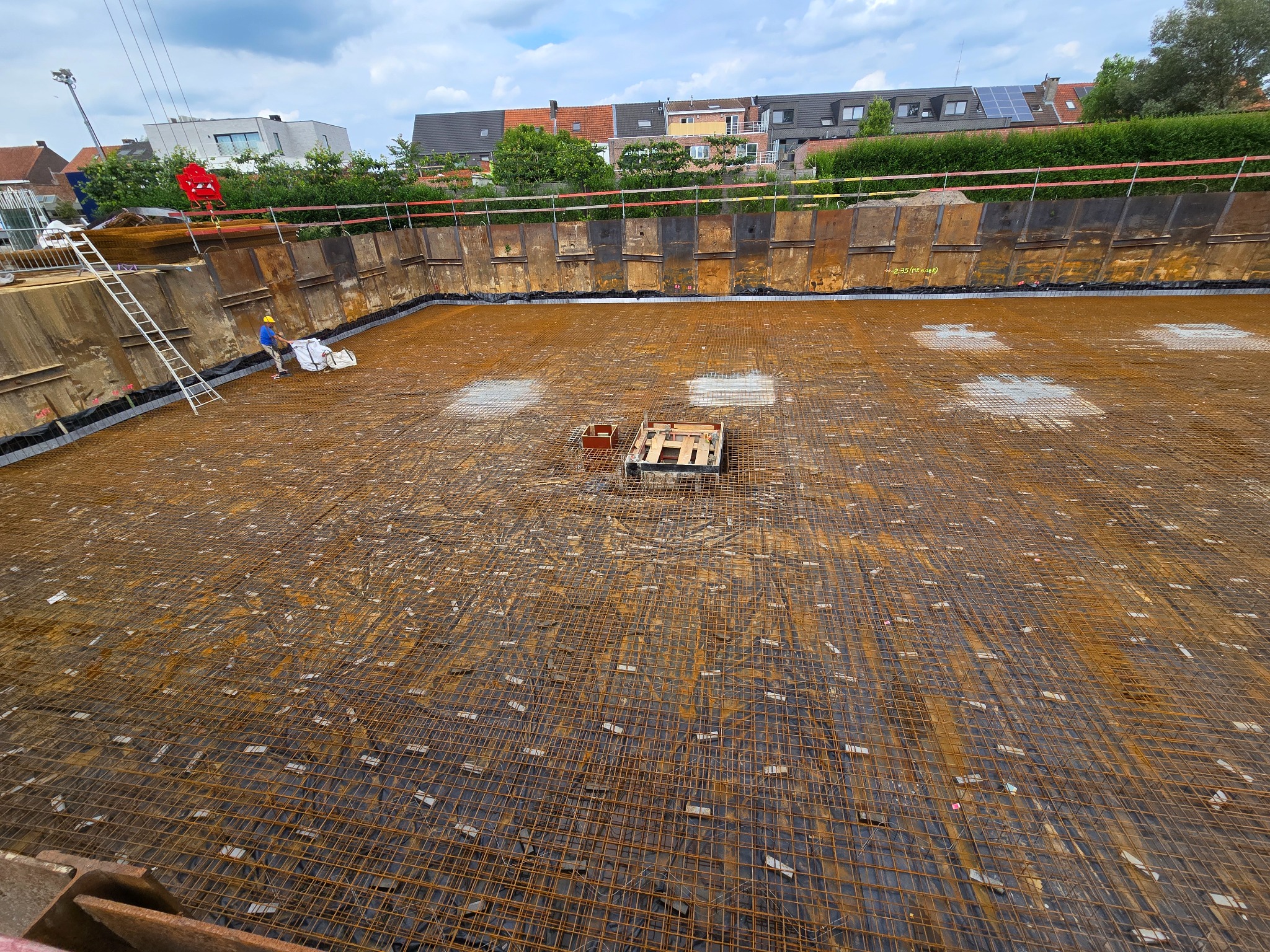 Szeroki wykop zabezpieczony metalowymi panelami, z ułożoną siatką zbrojeniową na czarnej folii, widoczne podpory i pracownik w kasku z workiem, częściowo pochmurne niebo.