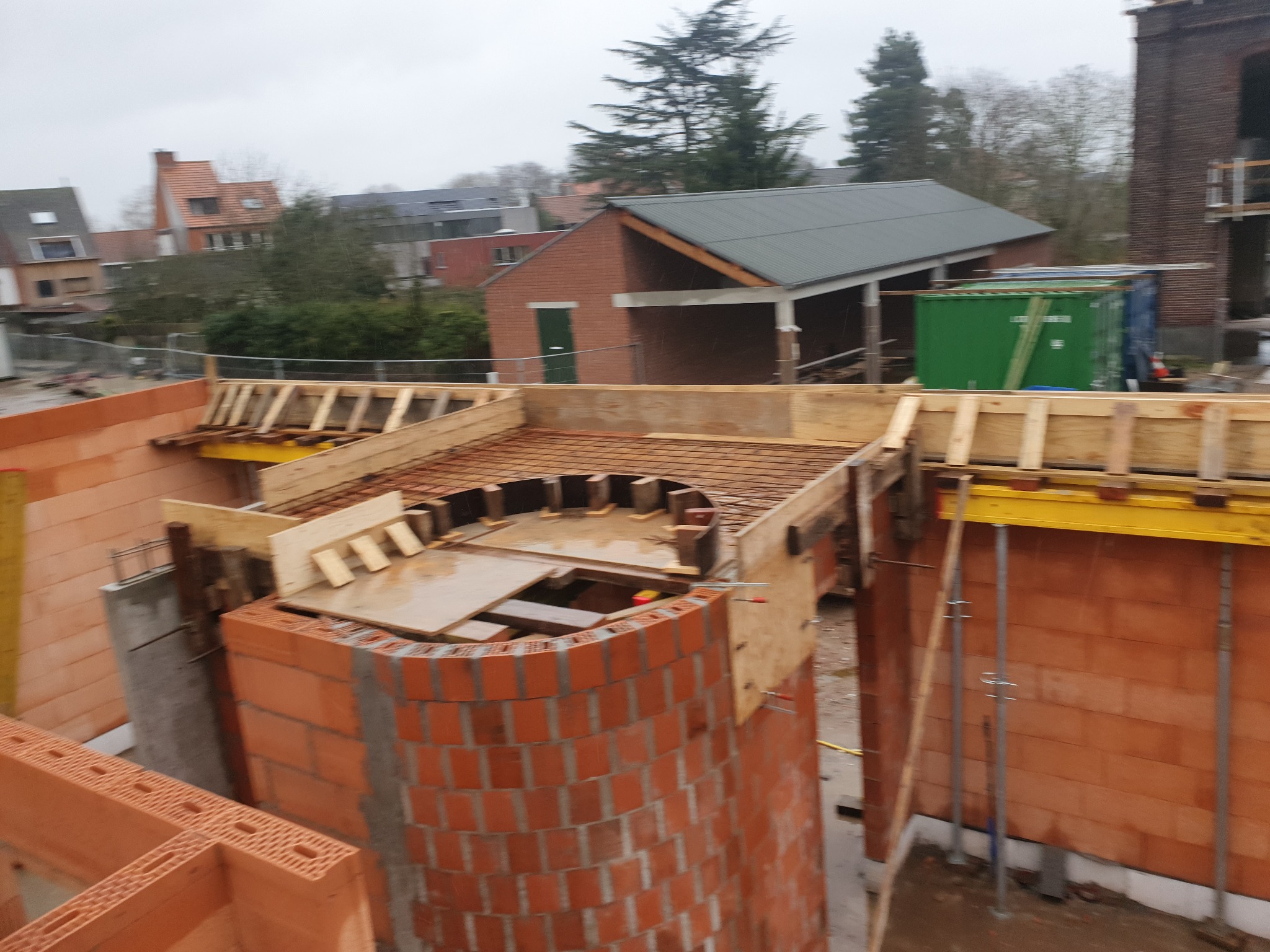 Budowa fundamentów z czerwonej cegły, widoczne szalunki i zbrojenia, w tle zabudowa mieszkalna i pochmurne niebo.