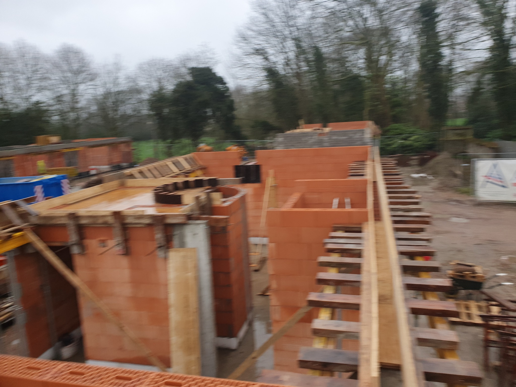 Budowa fundamentów z czerwonej cegły, widoczne drewniane szalunki i podpory, tło z drzewami w pochmurny dzień.