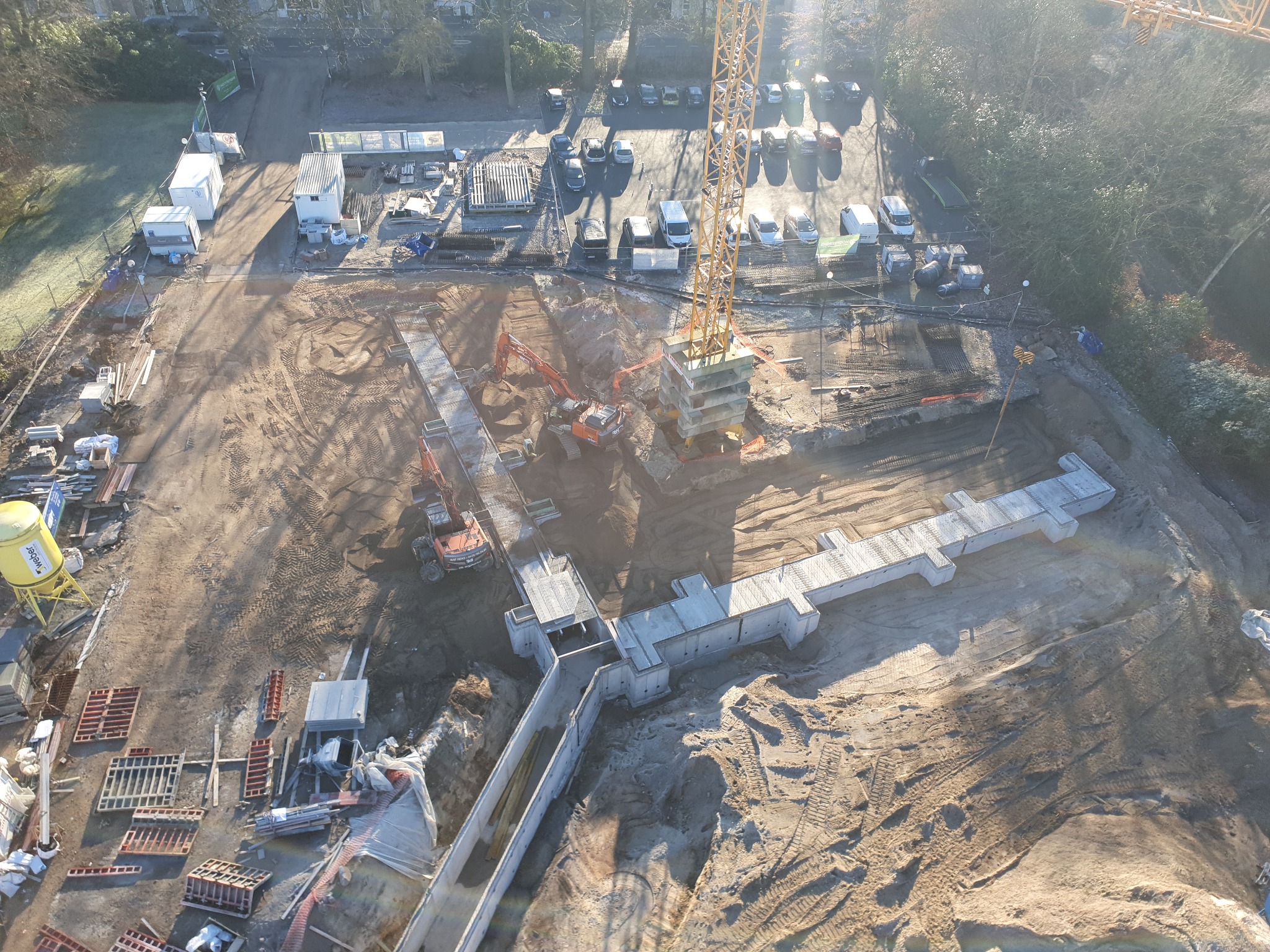 Widok z góry na plac budowy z wykopami, fundamentami w kształcie krzyża, koparkami i dźwigiem, w tle drzewa i parking.