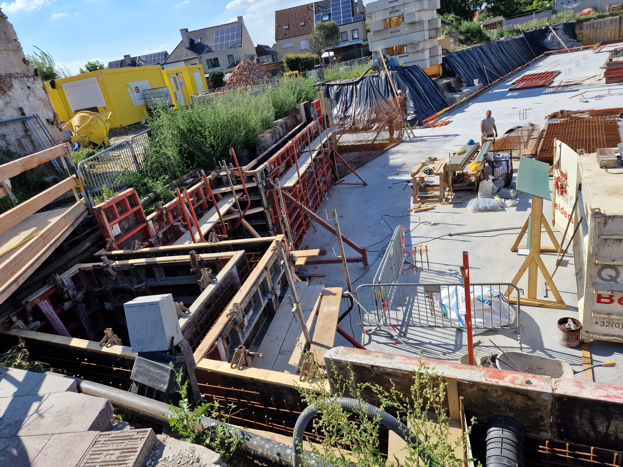 Budowa fundamentów: Szalunki zbrojeniowe przygotowane do zalewania betonem, widoczne pręty zbrojeniowe, teren budowy z kontenerami i materiałami budowlanymi w tle, na placu budowy stoi pracownik...