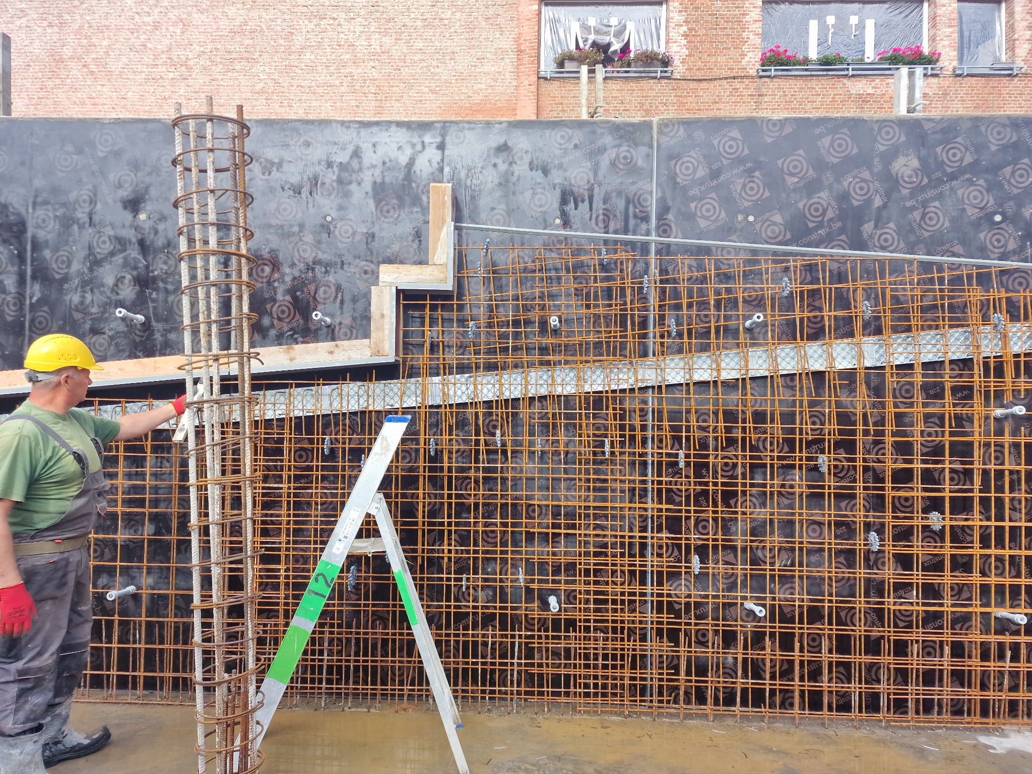 Pracownik w kasku i kombinezonie, z czerwoną rękawicą, przy zbrojeniu pionowego słupa na tle siatki zbrojeniowej ściany i drabiny aluminiowej z zielonymi elementami, widok na budynek z cegły...