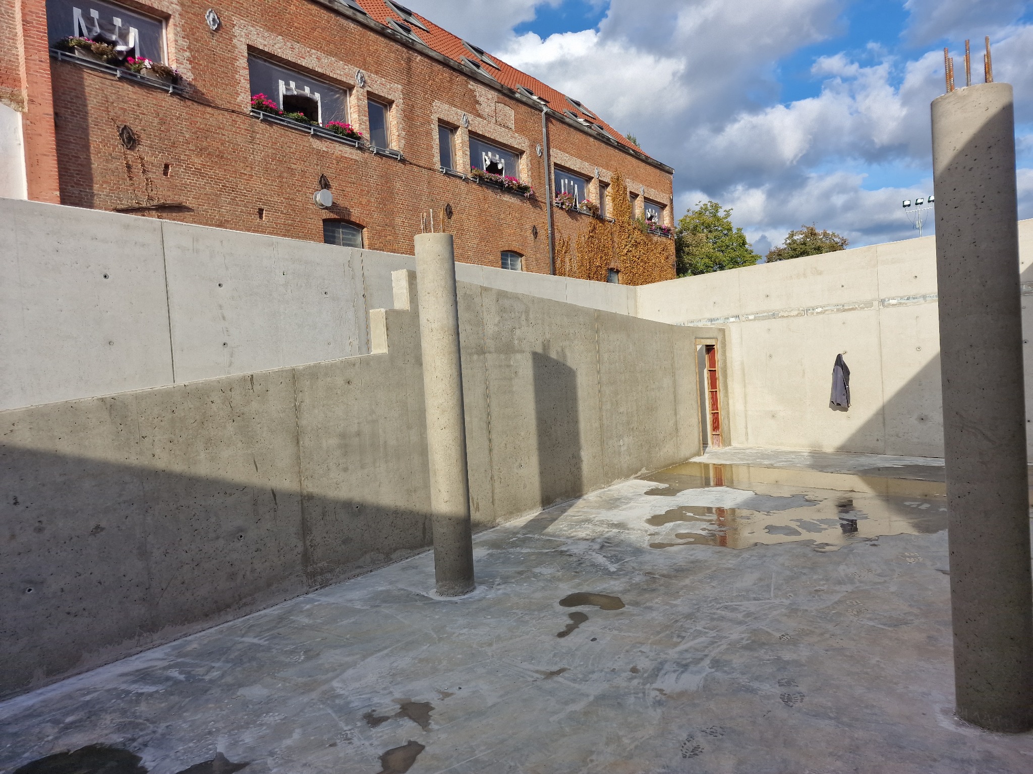 Surowy betonowy fundament budynku w trakcie budowy z widocznymi słupami zbrojeniowymi i śladami wody na posadzce, na tle ceglanego budynku z kwiatami w oknach.