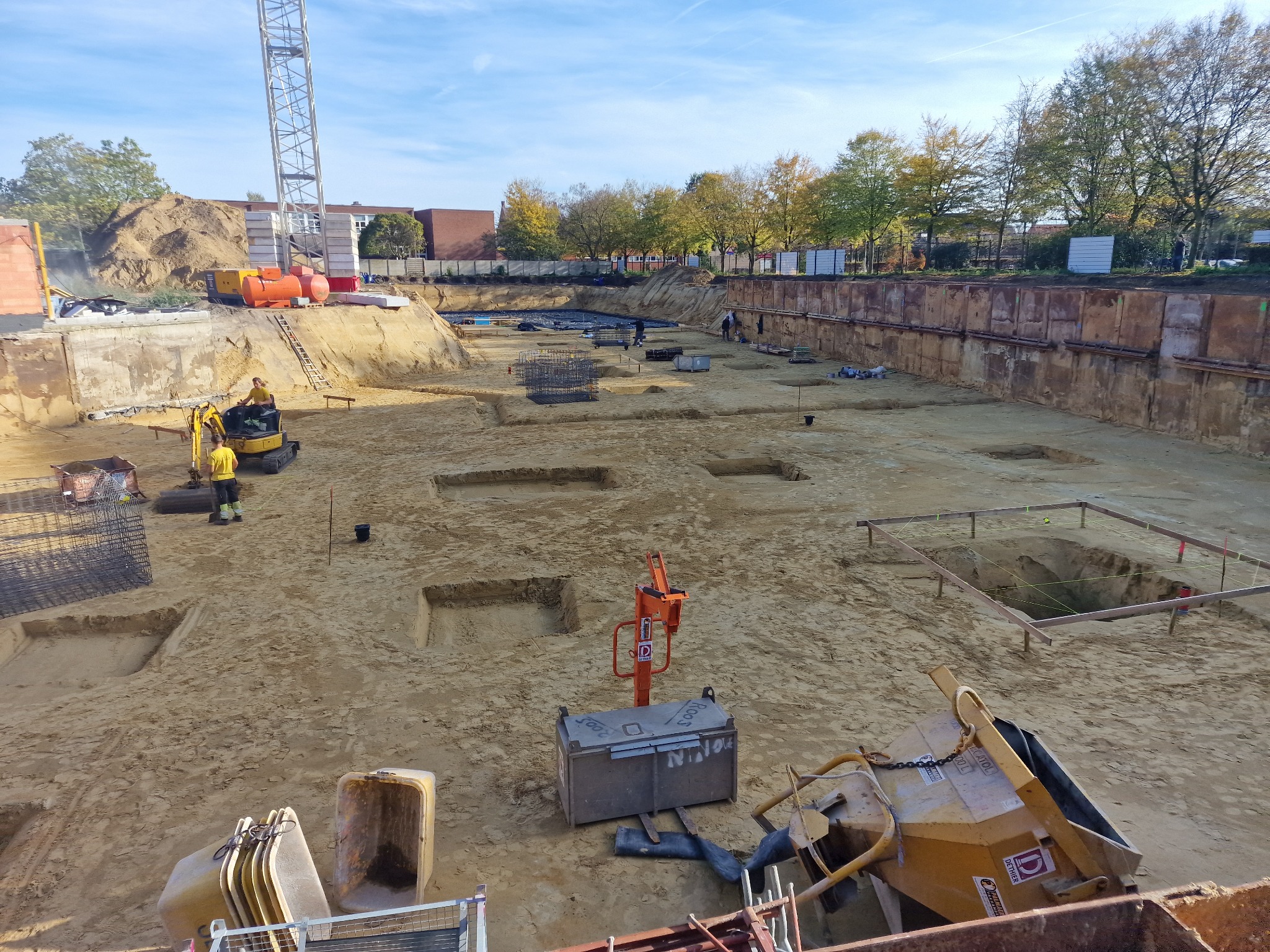 Szeroki wykop budowlany z zabezpieczonymi ścianami, przygotowane wykopy pod fundamenty, widoczny dźwig budowlany, mini koparka i pracownicy w oddali.
