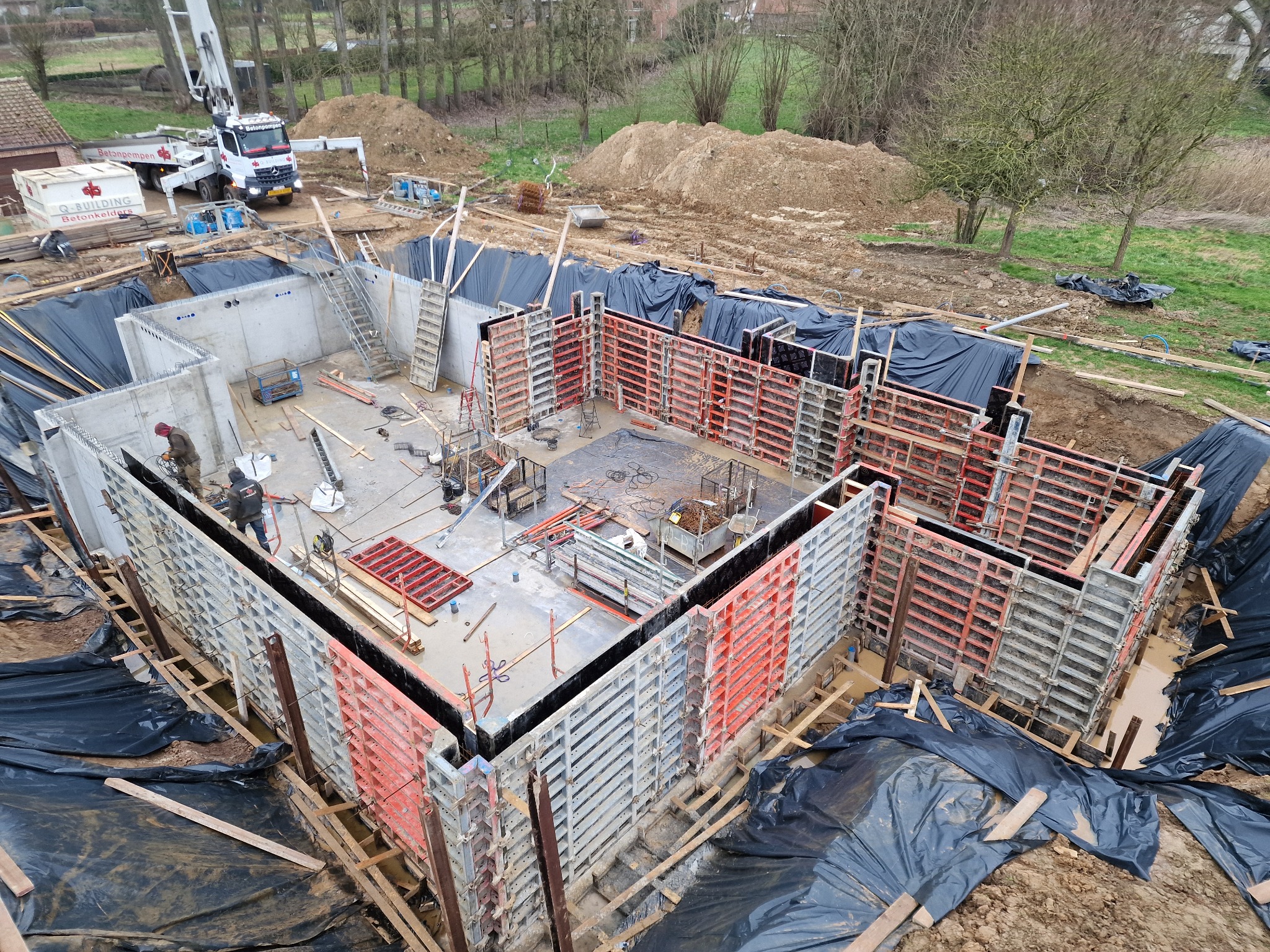 Budowa fundamentów: Widok z góry na częściowo zalany wykop z betonowymi ścianami szalunkowymi, zabezpieczony folią, z pracownikiem w tle i pompą do betonu na zewnątrz wykopu.