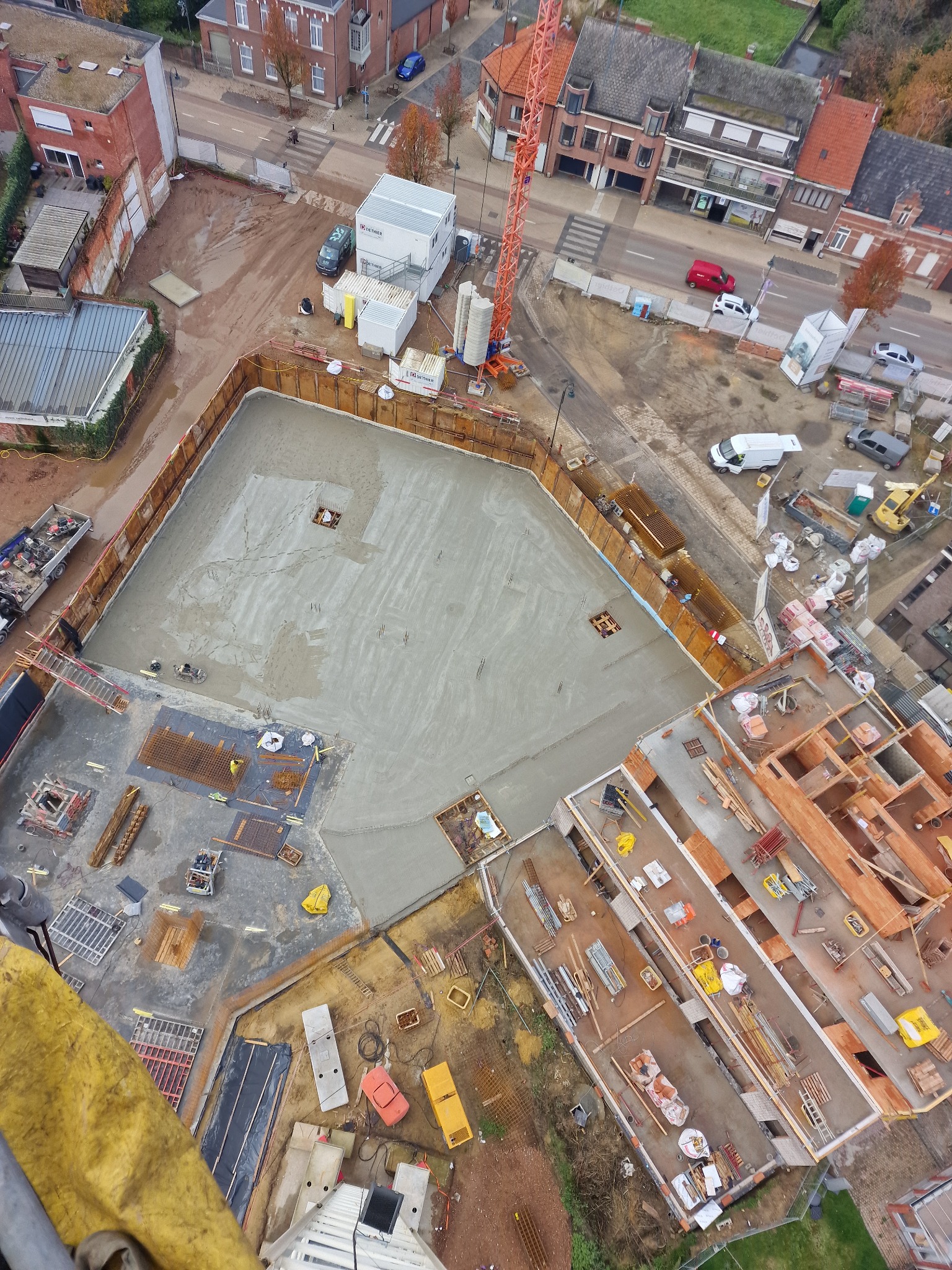 Widok z góry na plac budowy z wylanym betonem na fundamenty, widoczne szalunki, zbrojenia i maszyny budowlane, oraz częściowo ukończona konstrukcja budynku w tle.