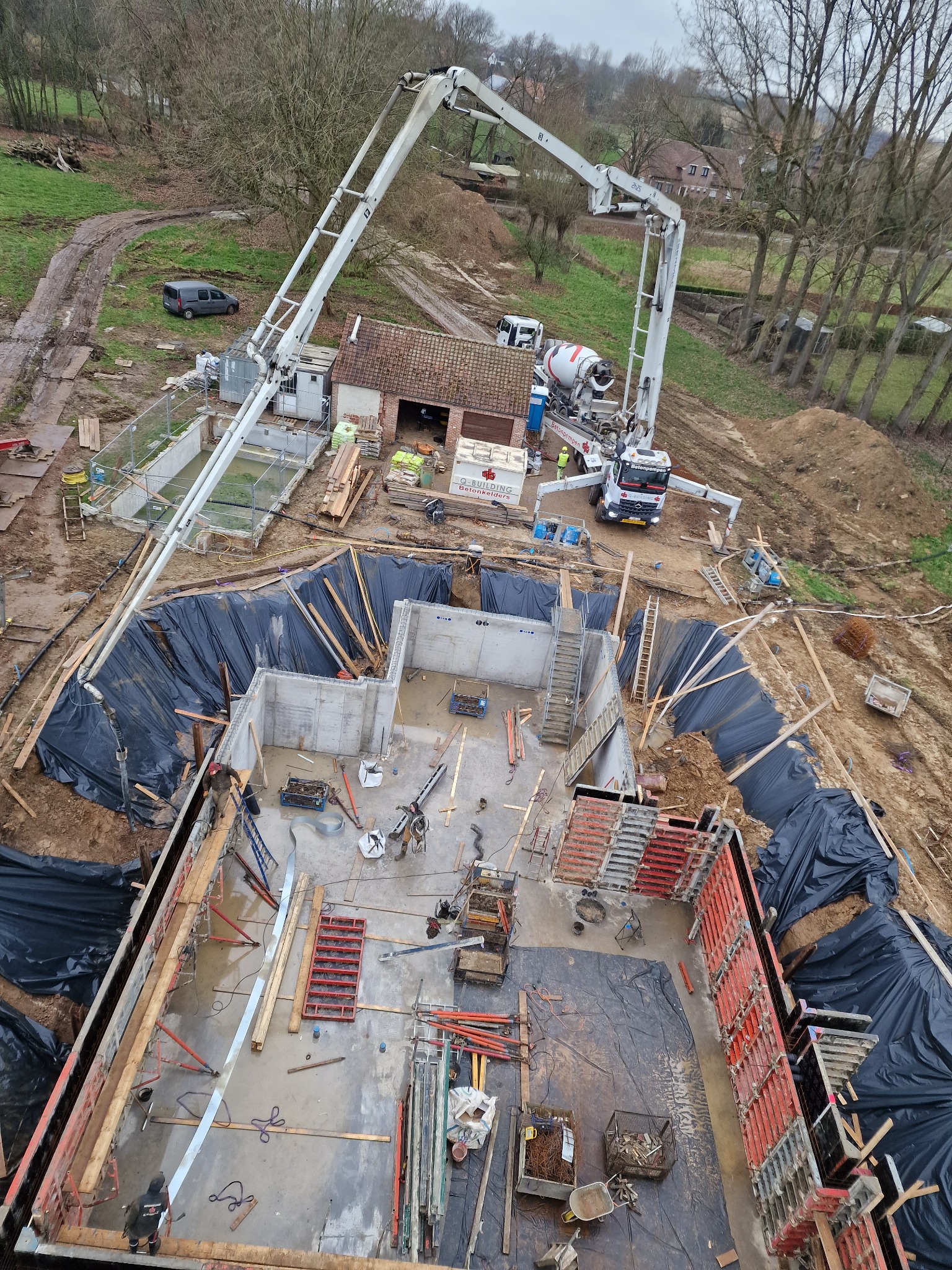 Widok z góry na plac budowy z wykopem pod fundamenty, częściowo zabetonowane ściany, szalunki, zbrojenia, pompę do betonu i betonomieszarkę w tle.