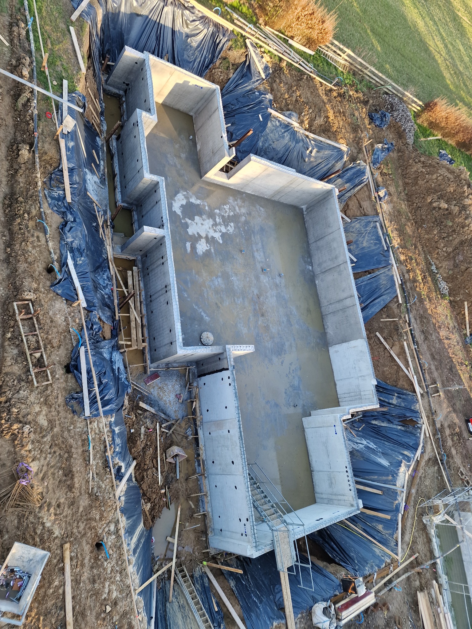 Widok z góry na fundamenty domu w trakcie budowy, z widoczną hydroizolacją z czarnej folii, deskowaniem i niewielką ilością wody wewnątrz, na tle ziemi i zieleni.