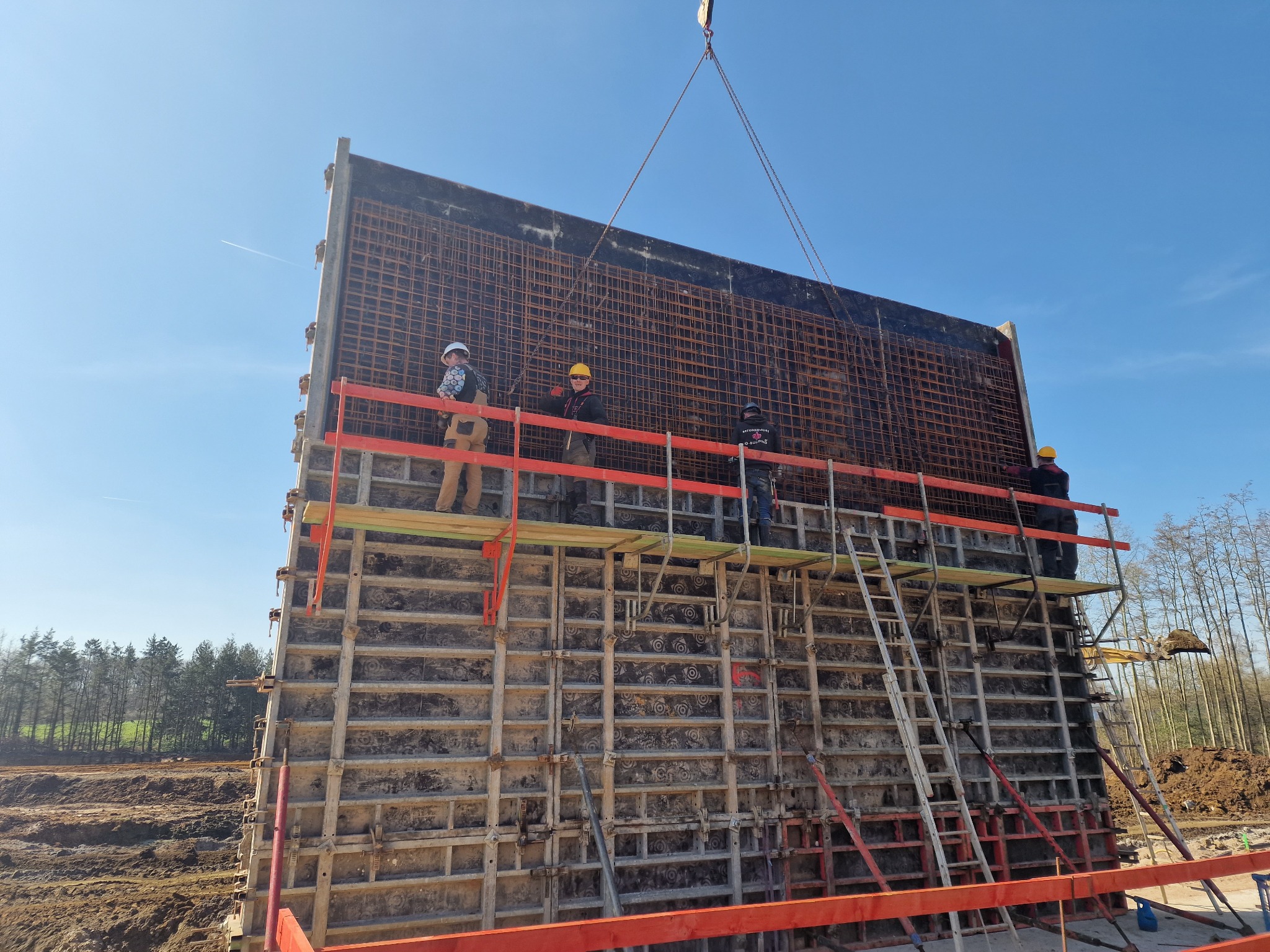 Montaż szalunku zbrojeniowego na budowie, pracownicy w kaskach na rusztowaniu, widoczne stalowe siatki zbrojeniowe i drewniane elementy konstrukcyjne, w tle błękitne niebo i drzewa.