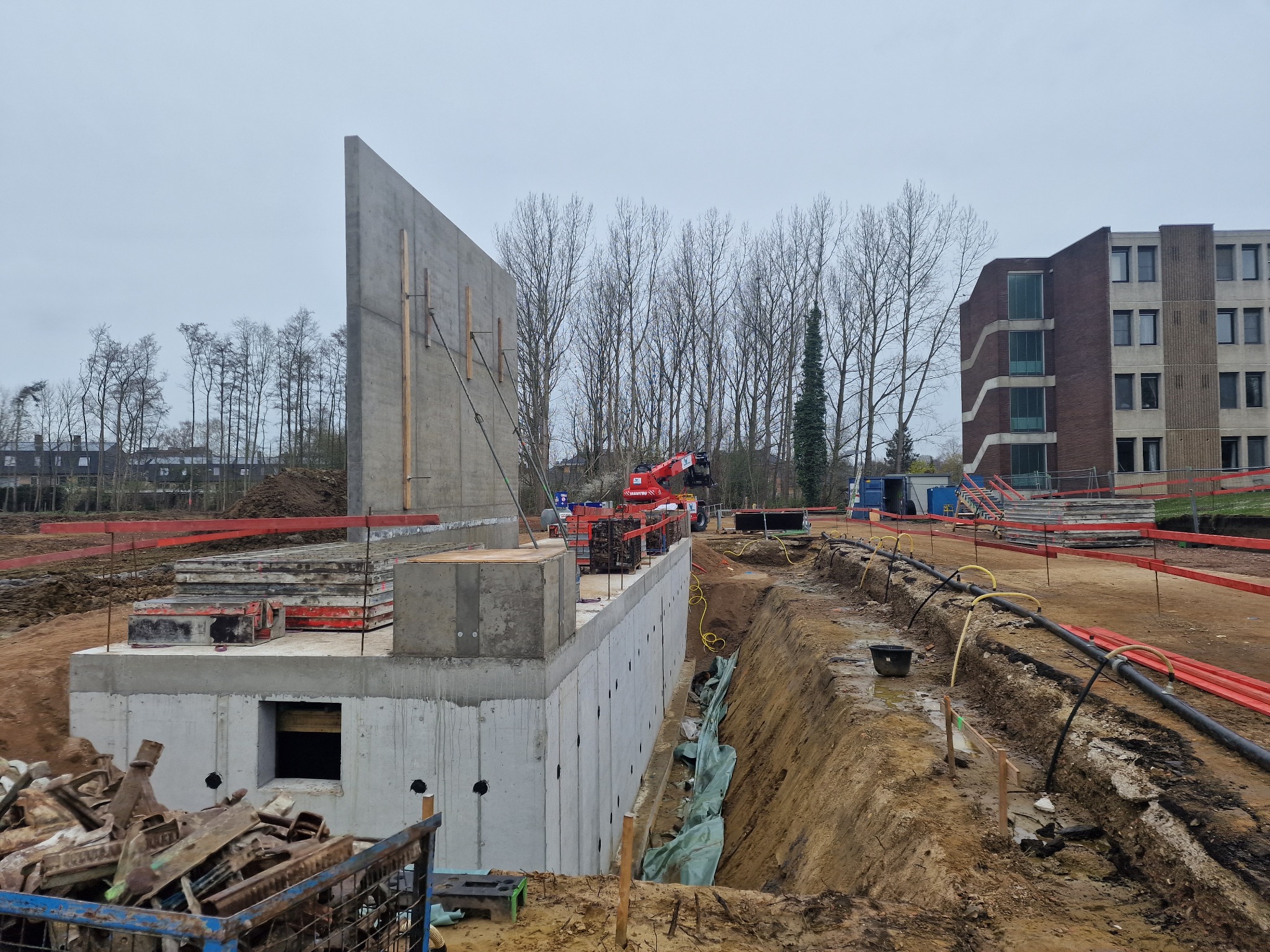 Wykop pod budynek z betonowymi ścianami fundamentowymi w trakcie budowy, widoczne szalunki, rury drenażowe i dźwig w tle.