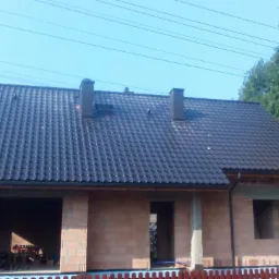 Niedokończony dom murowany z ciemną, błyszczącą dachówką, dwoma kominami i widocznymi rynnami, sfotografowany w słoneczny dzień.