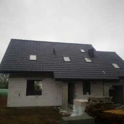 Dom w trakcie budowy z białego pustaka, z zamontowanym ciemnoszarym dachem z oknami połaciowymi i kominem, na tle pochmurnego nieba.