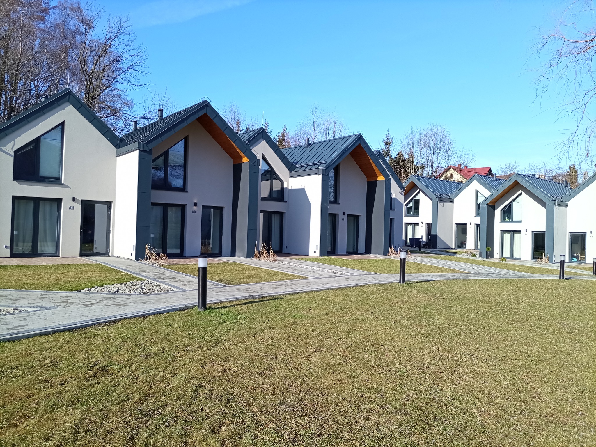 Szereg nowoczesnych domów jednorodzinnych z szarymi dachami i białymi elewacjami, widok z zewnątrz na tle błękitnego nieba i drzew bez liści.