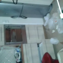 Zabrudzona podłoga w kuchni z resztkami jedzenia i śmieciami. Widoczne szafki kuchenne, okap i fragment krzesła. Niechlujny stan pomieszczenia przed sprzątaniem.