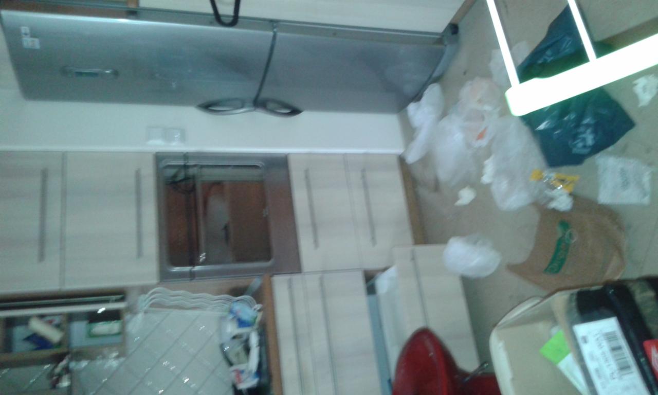 Zabrudzona podłoga w kuchni z resztkami jedzenia i śmieciami. Widoczne szafki kuchenne, okap i fragment krzesła. Niechlujny stan pomieszczenia przed sprzątaniem.