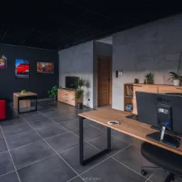 Nowoczesna aranżacja wnętrza poczekalni warsztatu samochodowego z czerwonym narożnikiem, biurkiem z monitorem LG i dekoracyjnymi obrazami samochodów na ścianie.