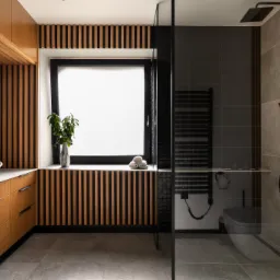 Nowoczesna łazienka z drewnianymi lamelami, owalną umywalką nablatową, czarną armaturą i kabiną prysznicową typu walk-in z czarnym szkłem.