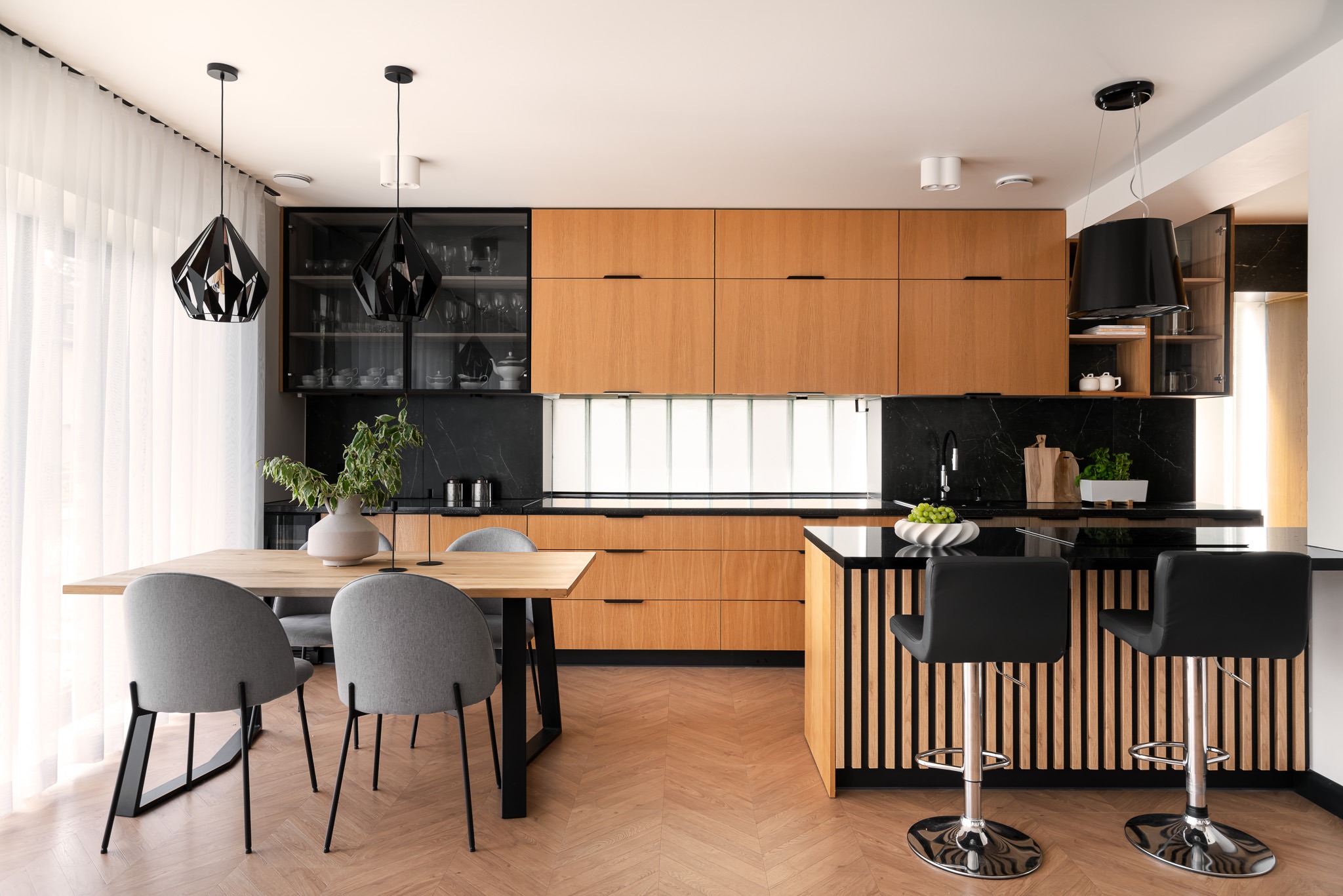 Nowoczesna kuchnia z drewnianymi szafkami, czarnym blatem i wyspą, oświetlona geometrycznymi lampami wiszącymi, uzupełniona stołem z szarymi krzesłami w Szczecinie.