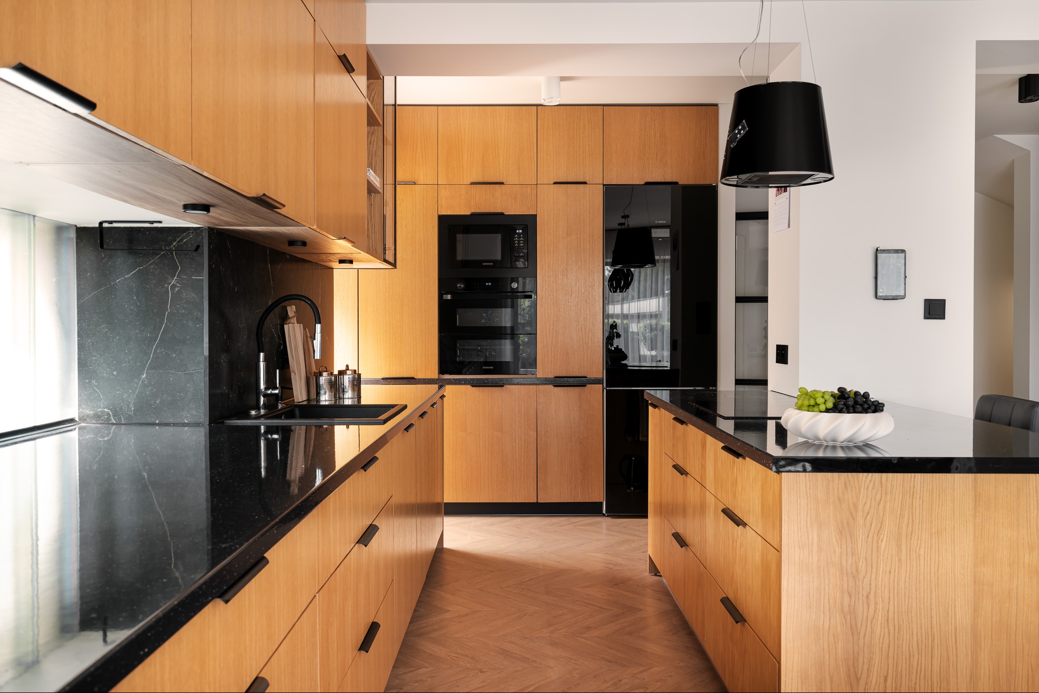 Nowoczesna kuchnia z drewnianymi szafkami, czarnym blatem i sprzętem AGD w zabudowie; na wyspie kuchennej misa z zielonymi i czarnymi winogronami.