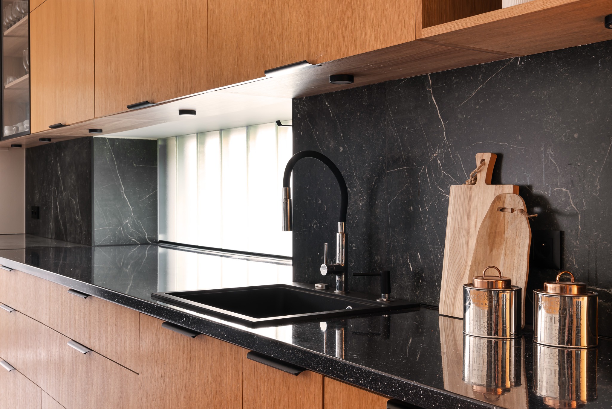 Nowoczesna kuchnia z drewnianymi szafkami, czarnym blatem z drobnymi białymi plamkami, czarnym zlewem i baterią z elastyczną wylewką, uzupełniona drewnianymi deskami do krojenia i metalowymi...