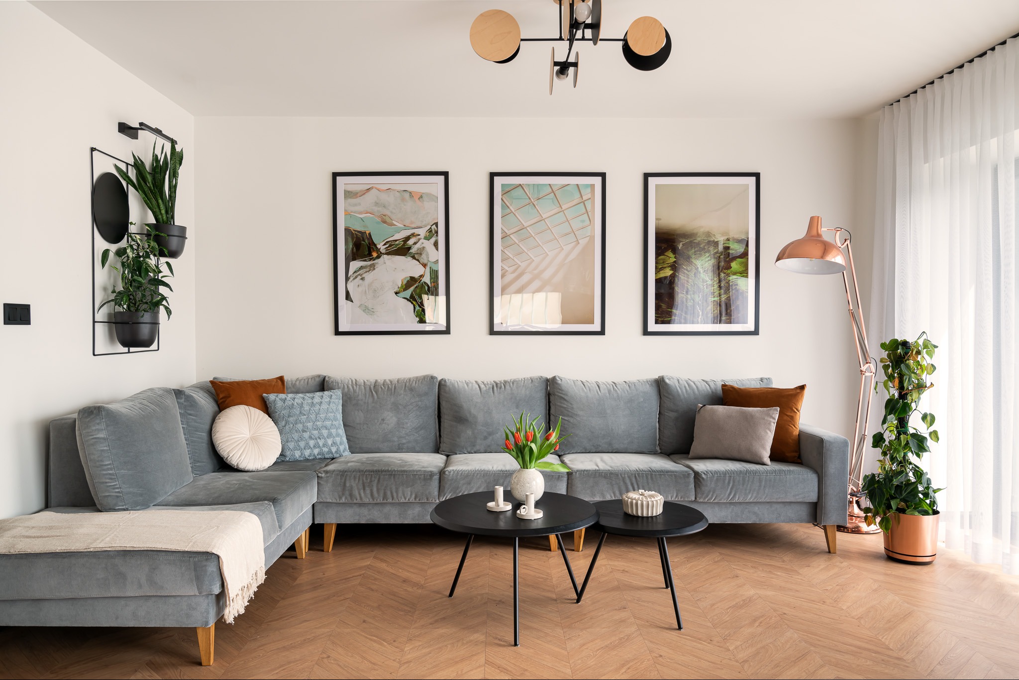 Jasny salon z szarą narożną sofą, trzema obrazami w czarnych ramach nad sofą, dwoma okrągłymi czarnymi stolikami kawowymi, lampą stojącą w kolorze miedzi i parkietem ułożonym w jodełkę.