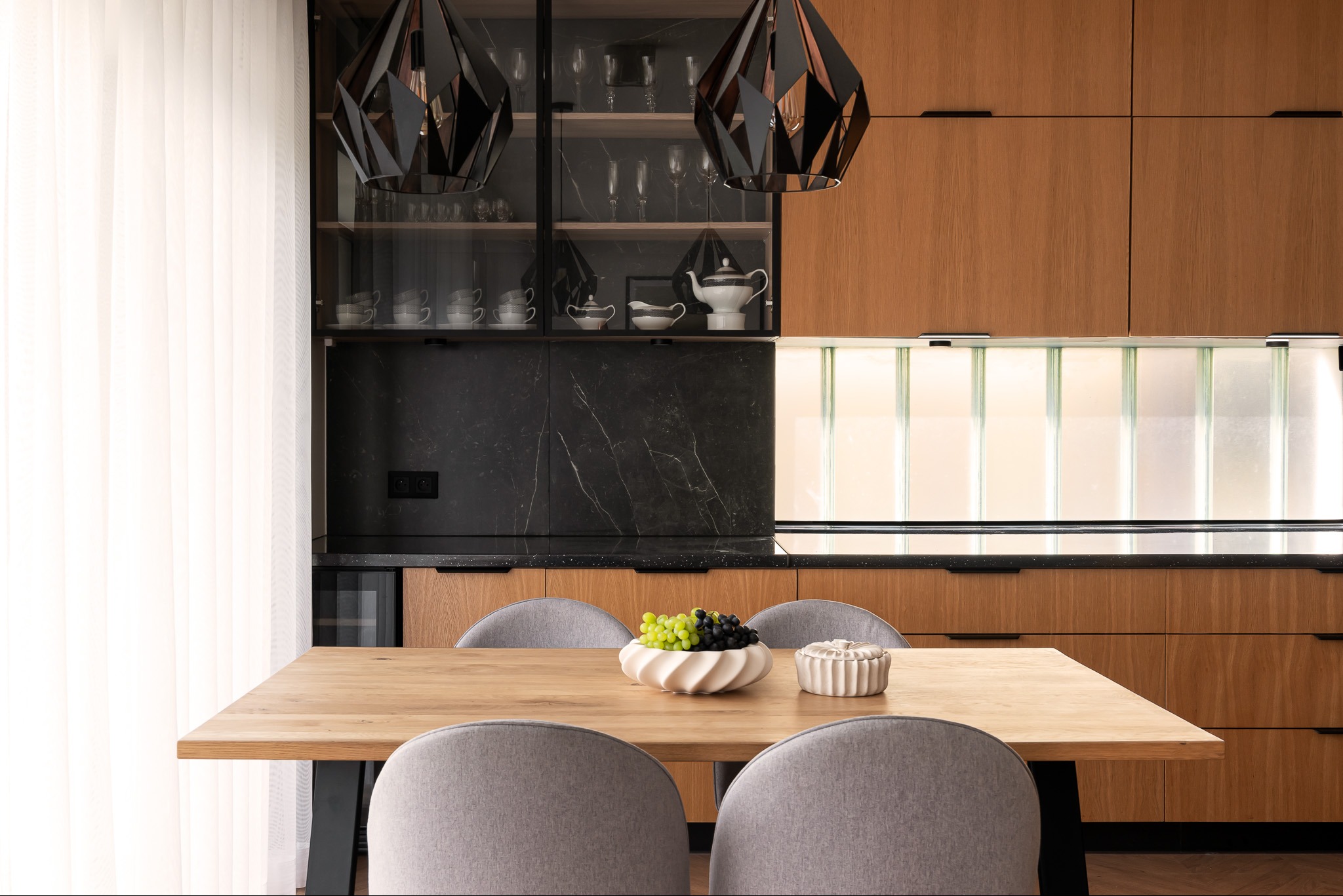 Nowoczesna kuchnia z drewnianymi szafkami, czarnym blatem, geometrycznymi lampami wiszącymi i drewnianym stołem z owocami w dekoracyjnej misie.
