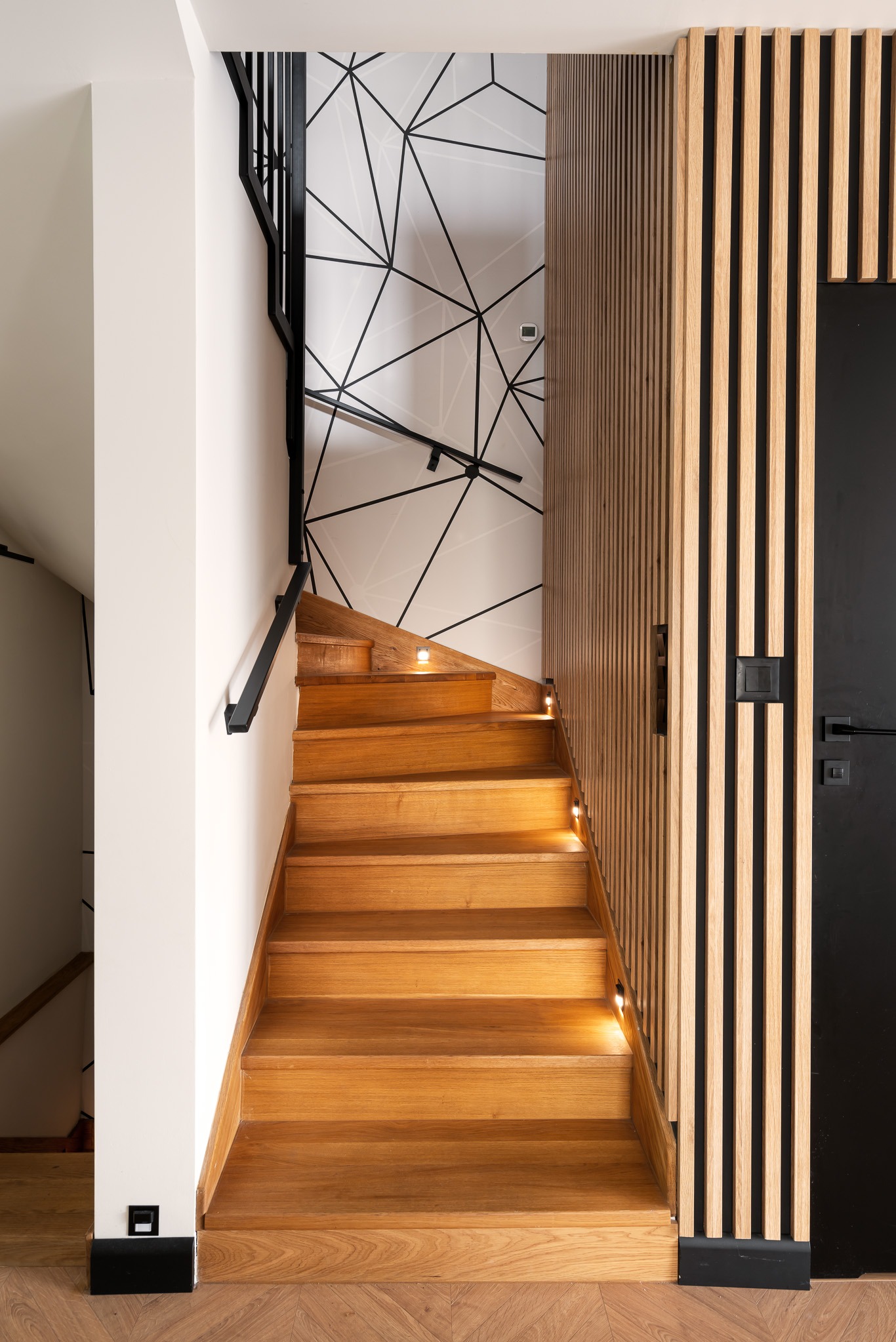Drewniane schody z podświetleniem LED na każdym stopniu, czarna balustrada, ściana z geometrycznym wzorem i drewniana boazeria z czarnymi drzwiami.