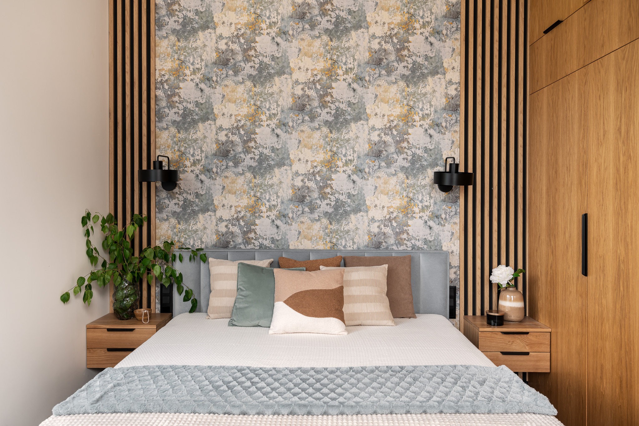 Nowoczesna sypialnia z łóżkiem, drewnianymi szafkami nocnymi i panelem dekoracyjnym z lameli na ścianie, wzorzysta tapeta w tle.