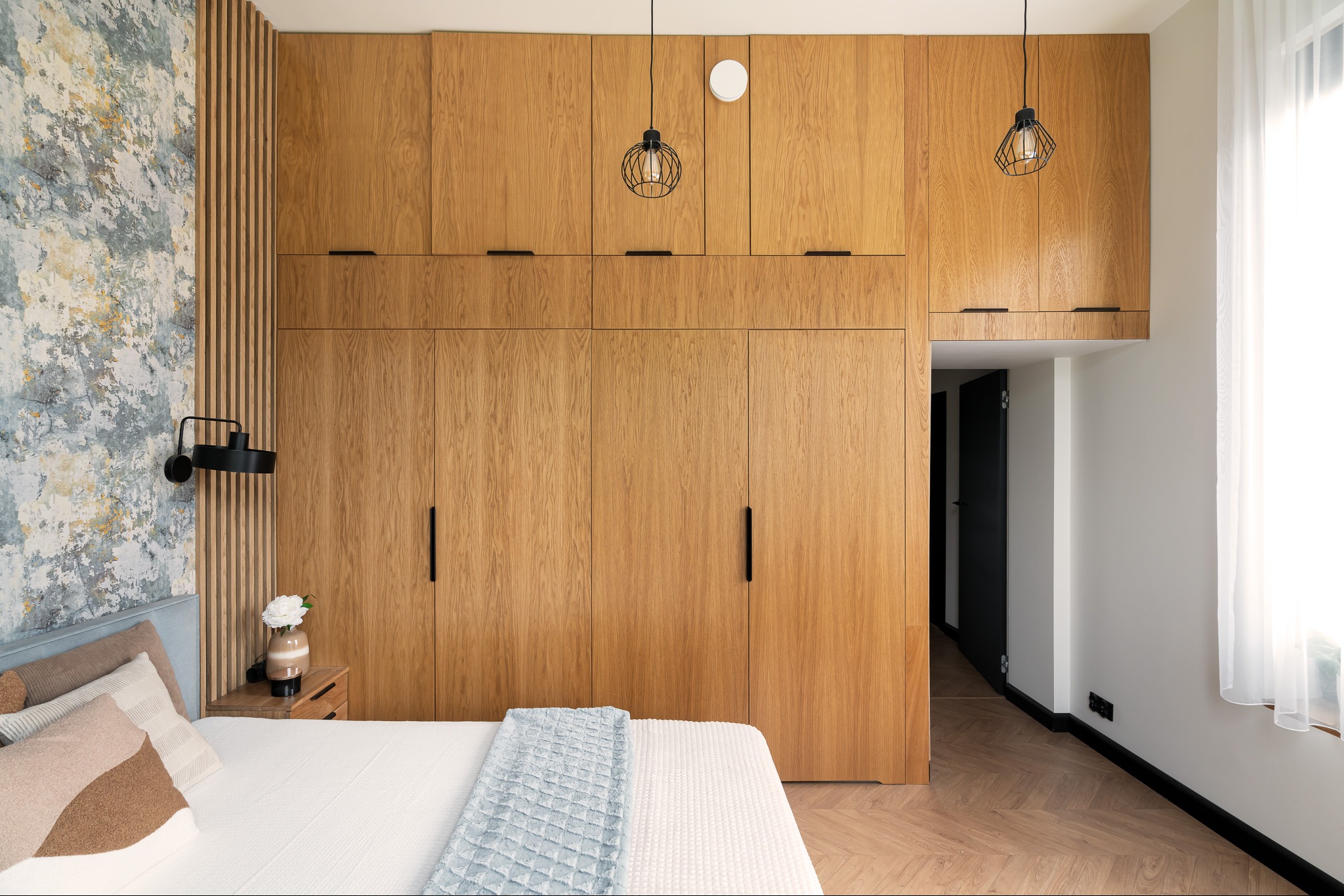 Nowoczesna sypialnia z drewnianą zabudową szaf od podłogi do sufitu, minimalistyczne czarne lampy wiszące, łóżko z dekoracyjnymi poduszkami i niebieskim pledem, parkiet ułożony w jodełkę.