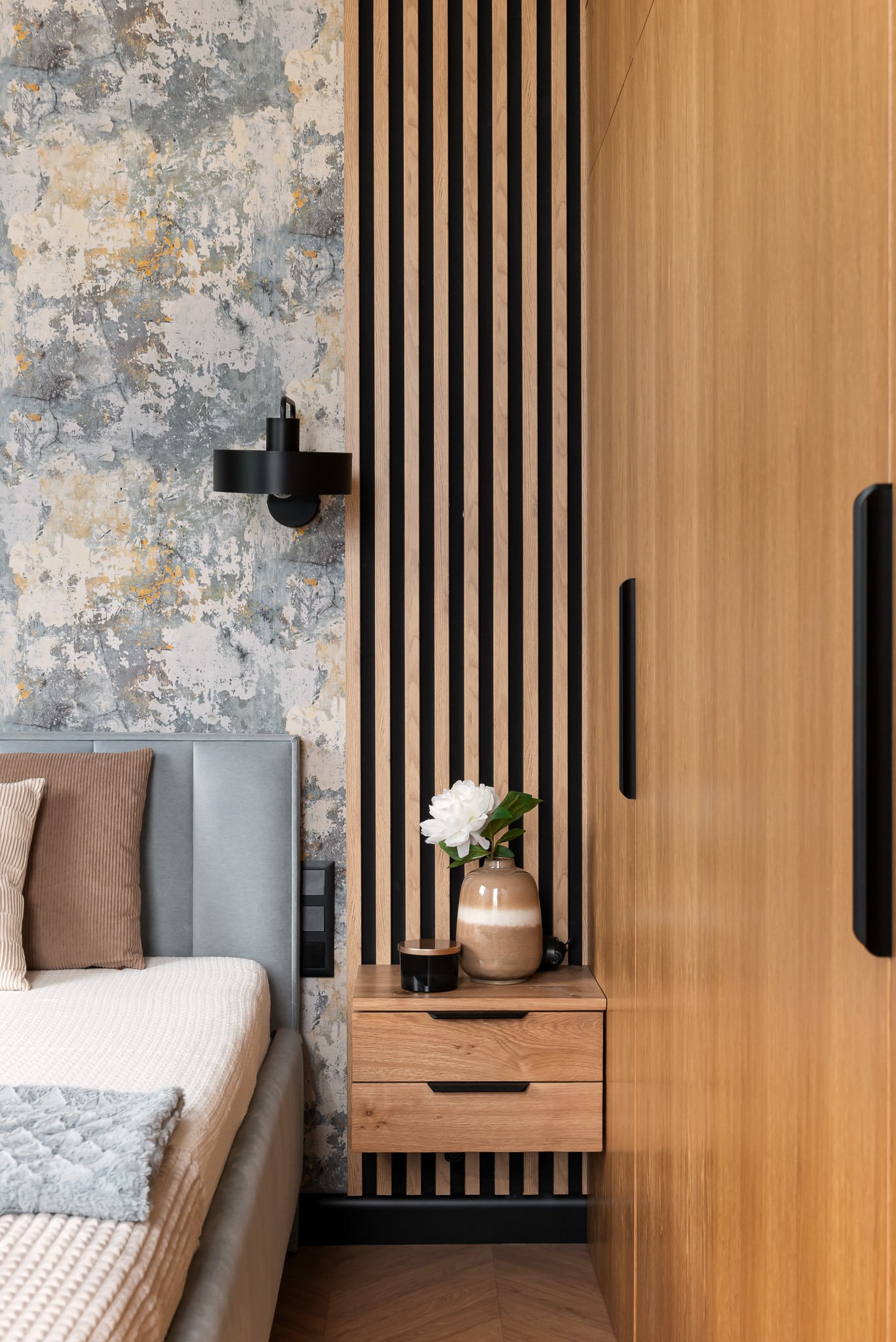 Fragment sypialni z łóżkiem, szafką nocną z drewnianymi lamelami i wazonem z białym kwiatem, na ścianie dekoracyjna faktura i czarny kinkiet