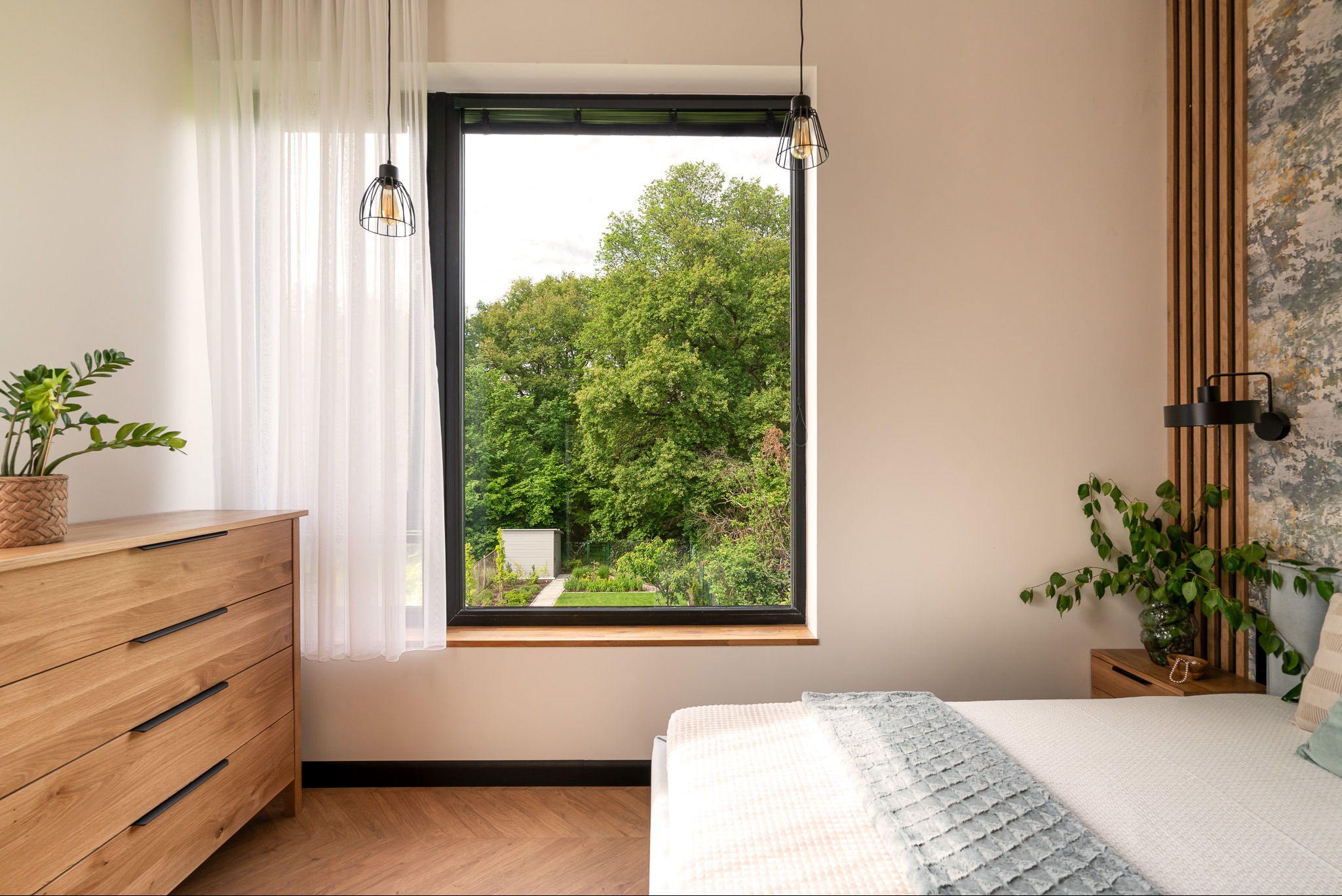 Jasna sypialnia z drewnianą komodą, łóżkiem z narzutą i widokiem na zielony ogród przez duże okno w czarnej ramie, uzupełniona lampami wiszącymi w stylu industrialnym.