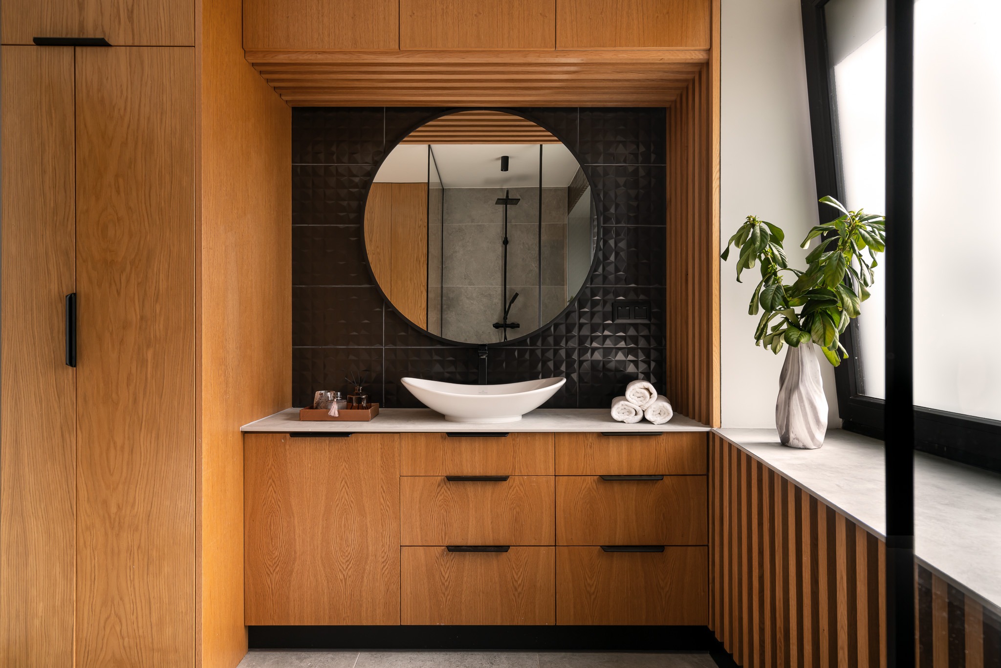 Nowoczesna łazienka z drewnianymi szafkami, okrągłym lustrem odbijającym kabinę prysznicową i umywalką w kształcie łodzi na blacie z marmuru.