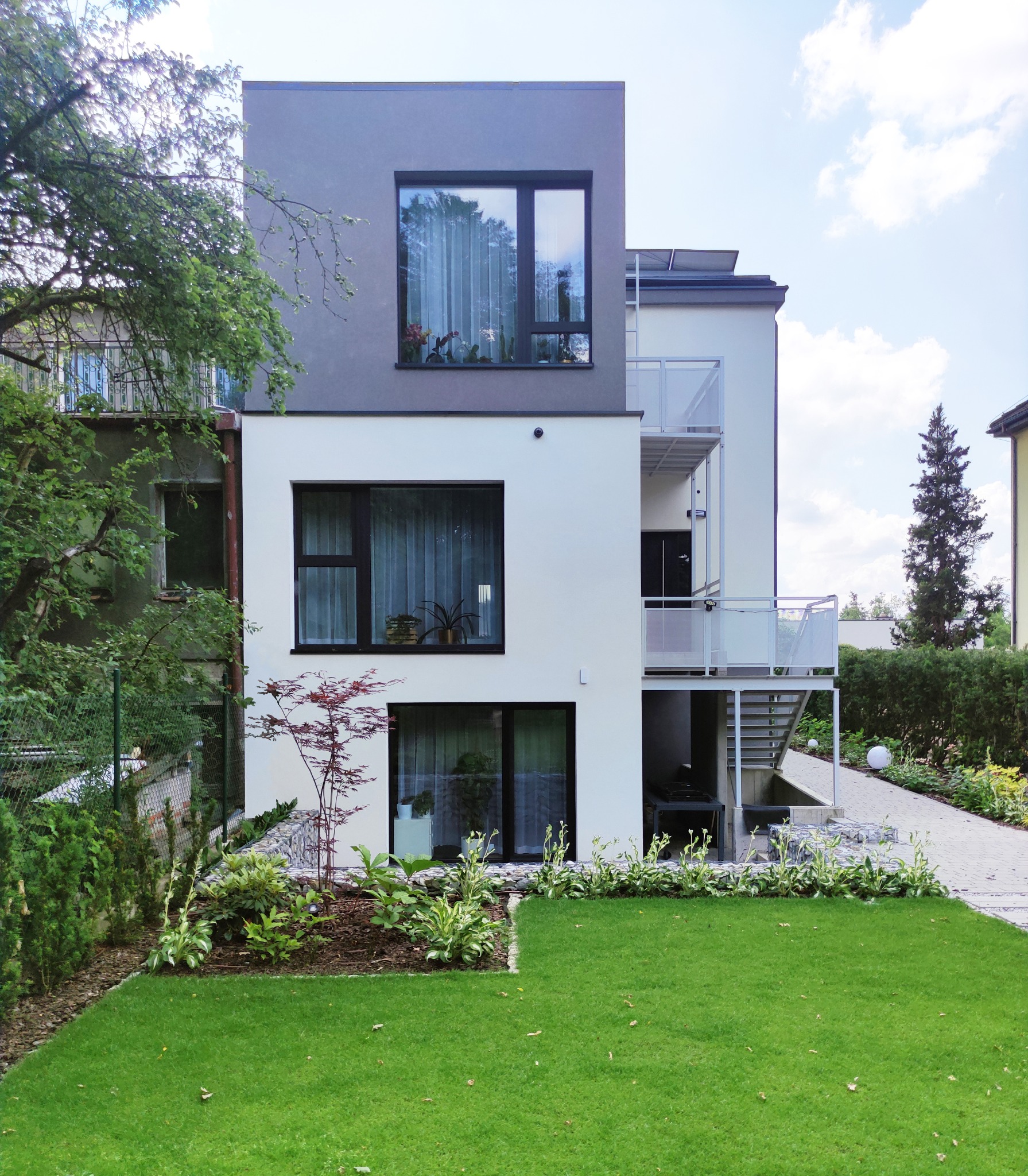 Nowoczesny dom z białymi ścianami i szarym górnym piętrem, z balkonami z białymi balustradami, otoczony zielonym trawnikiem i roślinnością ogrodową, widok z zewnątrz w słoneczny dzień.